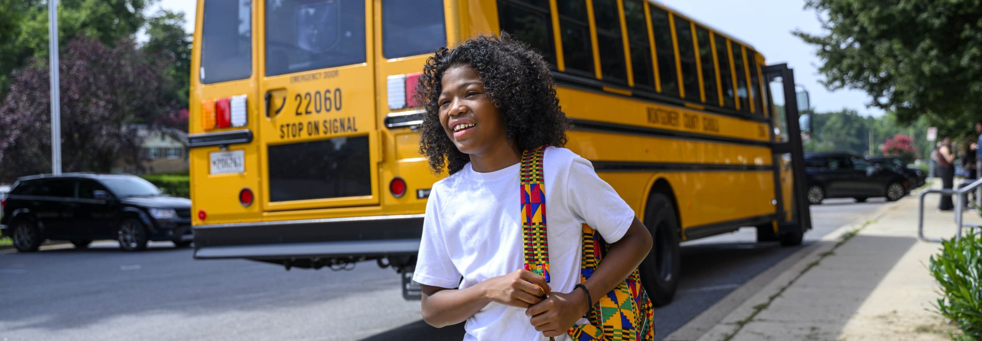 A smiling student with a colorful backpack stepping off a yellow electric school bus